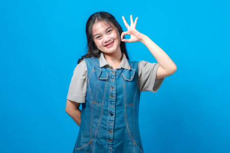 Cheerful young Asian woman smiling and showing an OK hand gesture, posing in front of a vibrant blue background, wearing casual denim and grey outfit.の写真素材