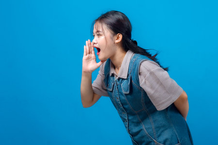 Side view of young Asian woman shouting with hand near mouth, showing calling gesture. Isolated on vibrant blue background, perfect for ads, communication, and marketing visuals.の写真素材