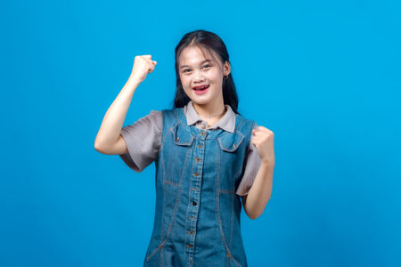 Cheerful young Asian woman smiling confidently while raising her fists in a gesture of success and motivation, standing against a solid blue background.の写真素材