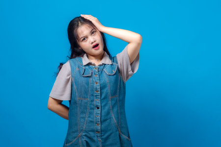 A young Asian woman poses in front of a solid blue background with a surprised and confused expression, touching her head while wearing a denim dress and casual shirt.の写真素材