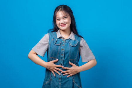 Young Asian woman smiling confidently with both hands on her stomach, expressing happiness, satisfaction, or digestive health. Studio portrait on blue background, front view.の写真素材