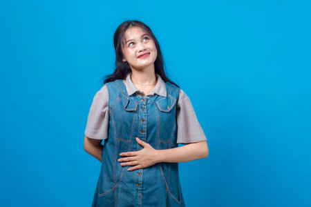 Young Asian woman looking up and smiling with hand on stomach, expressing hope, dreams, or positive imagination. Studio portrait isolated on blue background with copy space.の写真素材