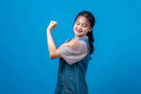 Confident young Asian woman smiling and flexing her bicep in a power pose against blue background. Her expression radiates empowerment, motivation, strength, and self-assurance.の写真素材