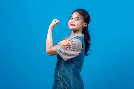 Confident young Asian woman smiling and flexing her bicep in a power pose against blue background. Her expression radiates empowerment, motivation, strength, and self-assurance.の写真素材