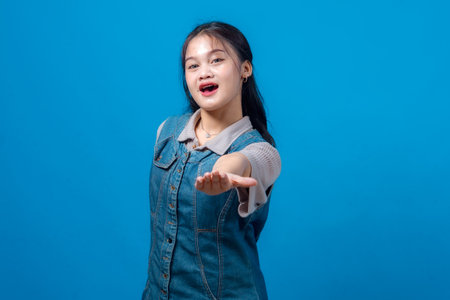 Cheerful young Asian woman gesturing to invite or welcome with one hand extended forward, smiling enthusiastically. Studio portrait against bright blue background, casual fashion.の写真素材