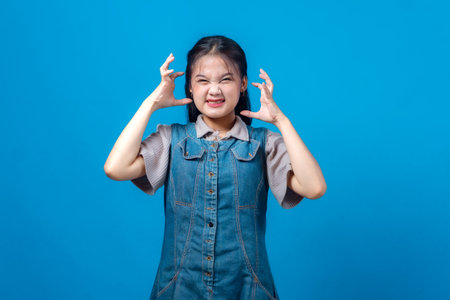 Frustrated young Asian woman making a stressed facial expression while clenching her hands near her head, wearing casual denim outfit on blue background. Mental pressure concept.の写真素材