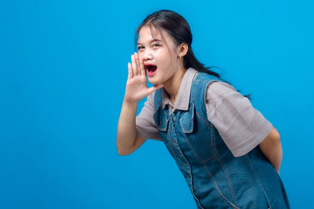 Young Asian woman shouting or calling out with hand near mouth, standing against blue background. She looks expressive, energetic, and enthusiastic.の写真素材
