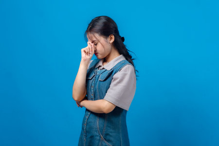 Upset young Asian woman closing her eyes and pinching the bridge of her nose, expressing stress or frustration. Studio portrait on blue background, suitable for emotional concept ads.の写真素材