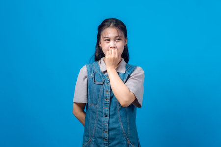 Portrait of young Asian woman expressing fear or anxiety with clenched fists near her face, looking upward. Studio shot on blue background, emotional reaction, denim outfit.の写真素材