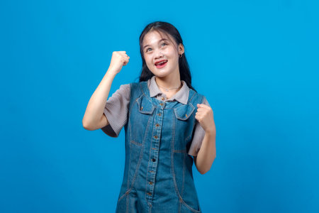 Joyful young Asian woman in denim dress raising her fists and looking up with excitement, expressing positivity, ambition, and hope against a clean blue studio background.の写真素材