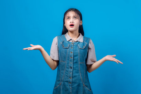 Confused young Asian woman raising both hands with a puzzled facial expression, looking upward. Studio shot on blue background, perfect for uncertainty or decision-making themes.の写真素材