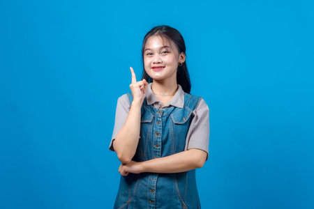 Cheerful young Asian woman smiling while raising index finger, suggesting an idea or solution, standing confidently in front of a plain blue background.の写真素材