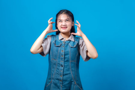 Annoyed young Asian woman looking up with a frustrated expression, clenching her hands in anger while standing against blue background. Concept of irritation, stress, and emotional tension.の写真素材
