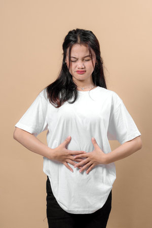 Young Asian woman in a white t-shirt clutching her stomach with a painful expression, indicating stomachache or digestive discomfort, isolated on beige background in studio.の写真素材