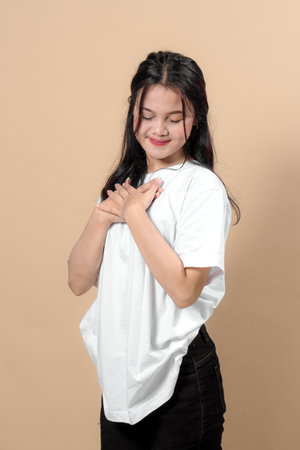 Young Asian woman in casual white t-shirt and black pants stands with closed eyes and hands crossed on chest, smiling softly, expressing gratitude, peace, and sincerity.の写真素材