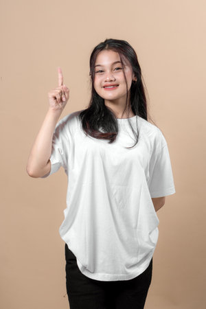 Smiling young Asian woman in a white t-shirt pointing up with her index finger, showing number one gesture, standing confidently against beige background in studio portrait.の写真素材
