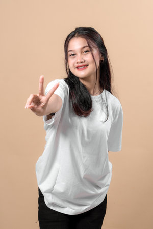 Young Asian woman smiling brightly while extending her arm forward showing a peace sign. She wears a plain white t-shirt and poses confidently against a soft beige background.の写真素材