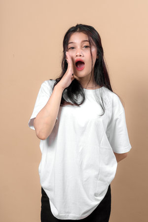 Young Asian woman shouting or calling out with hand near mouth, wearing a white t-shirt and standing against beige background, conveying urgency or spreading important news.の写真素材