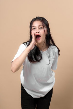 Asian woman in white t-shirt shouting with hand near mouth, leaning forward and making loud call gesture, expressing urgency or attention against plain beige background in studio.の写真素材