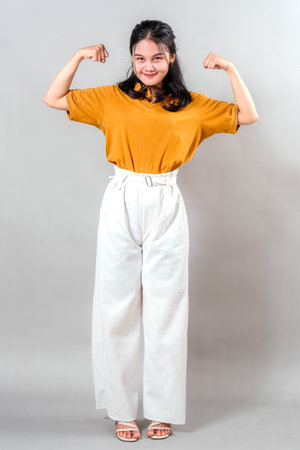 Smiling young Asian woman proudly flexing both arms in a strong pose, exuding confidence, charm, and determinationâideal for themes of empowerment, positivity, and motivation.の写真素材