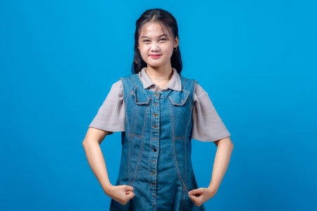 Confident young Asian woman showing a strong pose with clenched fists and slight smile, wearing a denim dress, standing against a vibrant blue background. Symbol of inner strength.の写真素材