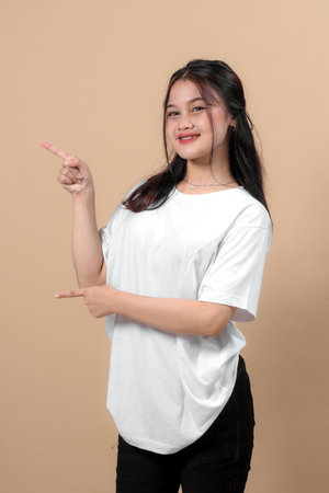 Happy young Asian woman in a white t-shirt pointing to the side with both hands while smiling, isolated on beige background. Perfect for promotional content and advertising mockups.の写真素材