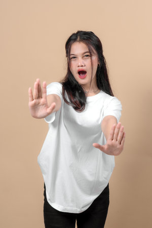 Young Asian woman raising both hands in a stop gesture with an intense, expressive face, conveying refusal or warning. Isolated on beige background, studio shot.の写真素材