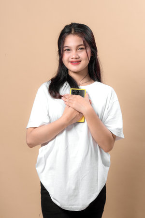 Young Asian woman smiling while holding a yellow smartphone close to her chest, wearing a casual...