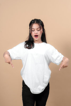 A young Asian woman in a white oversized t-shirt and black pants points downward with both hands, looking surprised and excited, on a plain beige background.の写真素材