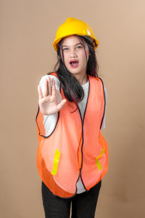 Asian woman in safety vest and yellow hard hat extending her hand forward in a "stop" gesture, showing a serious warning expression. Concept of caution, restriction, and safety.の写真素材
