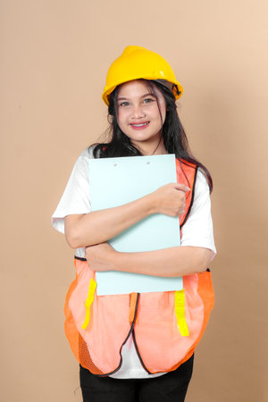 Confident young woman in construction attire, wearing an orange safety vest and yellow hard hat,...
