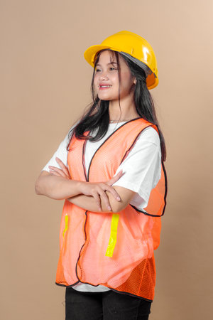 Confident young Asian female construction worker in safety gear, including a yellow hard hat and orange reflective vest, standing with arms crossed, smiling.の写真素材