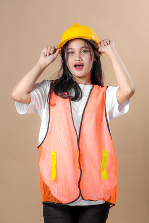 Young Asian female worker wearing a white t-shirt, orange safety vest, and yellow hard hat poses confidently with a cheerful expression on a neutral background.の写真素材