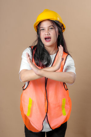 Young Asian woman in orange safety vest and yellow hard hat making an "X" hand gesture in front of her chest, showing refusal or prohibition with a serious facial expression.の写真素材