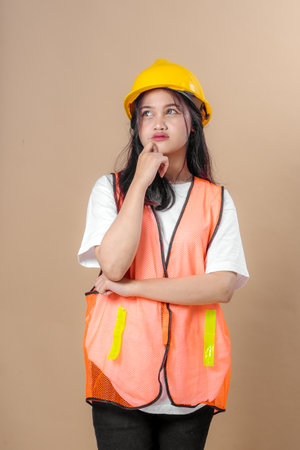 Portrait of a young female construction worker wearing a yellow hard hat and orange safety vest, standing with crossed arms and a thinking expression, isolated on beige background.の写真素材
