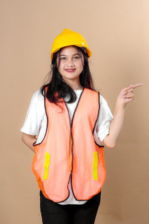 Smiling young woman in a yellow hard hat and orange safety vest confidently pointing to the side, symbolizing guidance, direction, or instruction in a professional construction setting.の写真素材