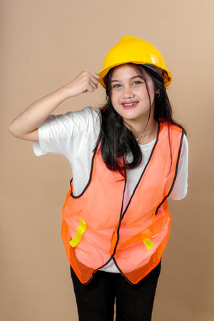Young Asian woman in orange safety vest and yellow hard hat posing confidently with a cheerful smile and a raised fist gesture, symbolizing motivation, strength, and work dedication.の写真素材