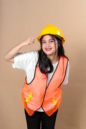 Young Asian woman in orange safety vest and yellow hard hat posing confidently with a cheerful smile and a raised fist gesture, symbolizing motivation, strength, and work dedication.の写真素材