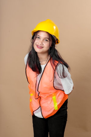 A cheerful young woman wearing a yellow hard hat and orange safety vest leans forward slightly and smiles brightly, posing confidently in a construction-themed outfit.の写真素材
