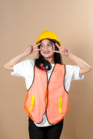 A young woman in a yellow hard hat and orange safety vest makes a double peace sign while smiling confidently, representing cheerful construction safety awareness on beige background.の写真素材