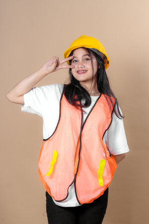 A cheerful young woman in a yellow hard hat and orange safety vest poses with a peace sign near her eye, symbolizing confidence, security, and a positive attitude in construction work.の写真素材
