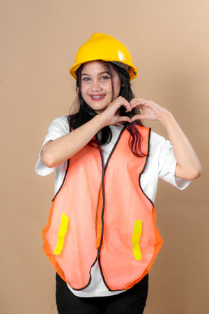 Cheerful young woman in a yellow construction helmet and orange safety vest smiling confidently while making a heart hand sign, representing care, safety, and positive workplace energy.の写真素材