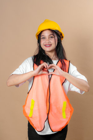 Young woman in a yellow hard hat and orange safety vest smiling warmly while making a heart gesture with her hands, symbolizing care and positivity at work.の写真素材