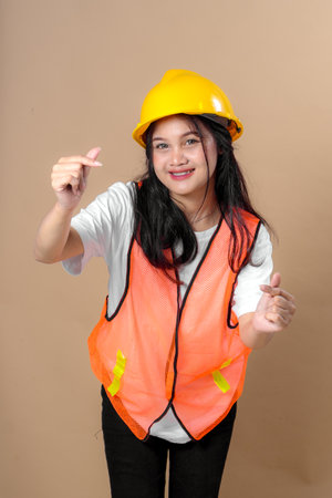 Smiling young woman in a yellow hard hat and orange safety vest making a finger heart gesture with both hands, expressing positivity at a worksite concept.の写真素材