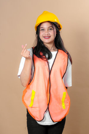 Smiling young woman wearing a yellow hard hat and orange safety vest showing Korean finger heart gesture, representing positivity, friendliness, and construction safety awareness.の写真素材