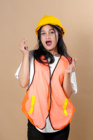 Young woman wearing a yellow hard hat and orange safety vest making a cheerful expression with double finger heart gestures, symbolizing friendliness, joy, and workplace positivity.の写真素材