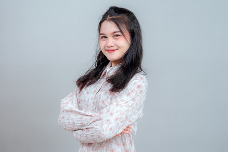 Portrait of a young Asian woman confidently smiling with arms crossed, expressing self-assurance and friendliness. Studio shot on plain background, perfect for business or lifestyle ads.の写真素材