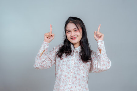 Young Asian woman smiling warmly while pointing both index fingers upward, expressing cheerful confidence and positive energy, ideal for advertising, guidance, or promotional content.の写真素材
