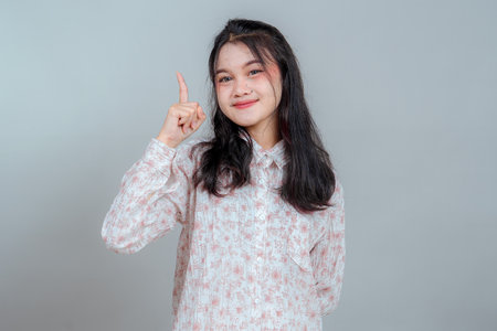 Confident young Asian woman with a friendly smile and braces pointing one finger up, suggesting an idea or solution. Positive facial expression, isolated on a plain background.の写真素材
