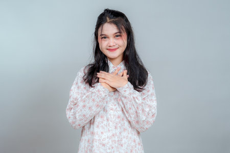 Young Asian woman smiling warmly with hands over heart, expressing sincere gratitude and kindness. Emotional portrait of thankfulness and affection in studio setting on gray background.の写真素材